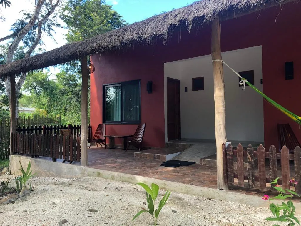 Porche de una cabaña de paredes rojizas, con techo de palapa y una hamaca